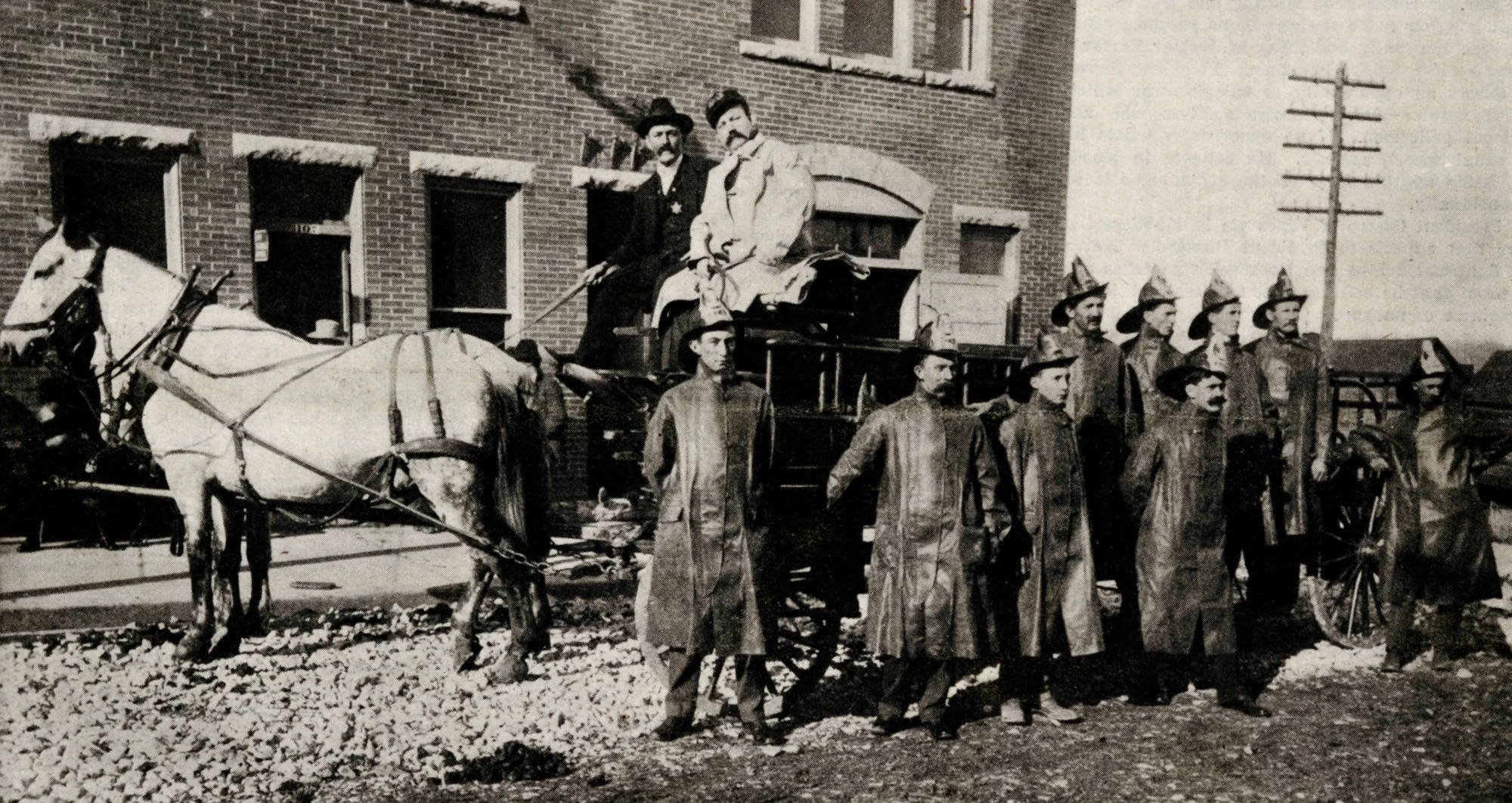 Columbia's early firefighters in long coats and helmets beside a horse-drawn fire wagon, with two white horses hitched at left